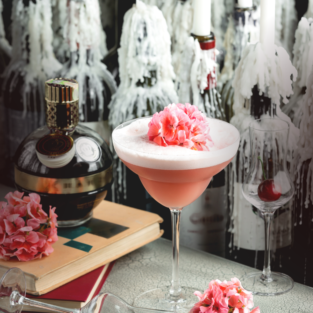 A stylish pink cocktail garnished with pink flowers, served in a coupe glass surrounded by decorative elements including vintage books, wax-covered wine bottles, and a cherry in a liqueur glass—evoking an elegant, Kate Spade-inspired party setting.