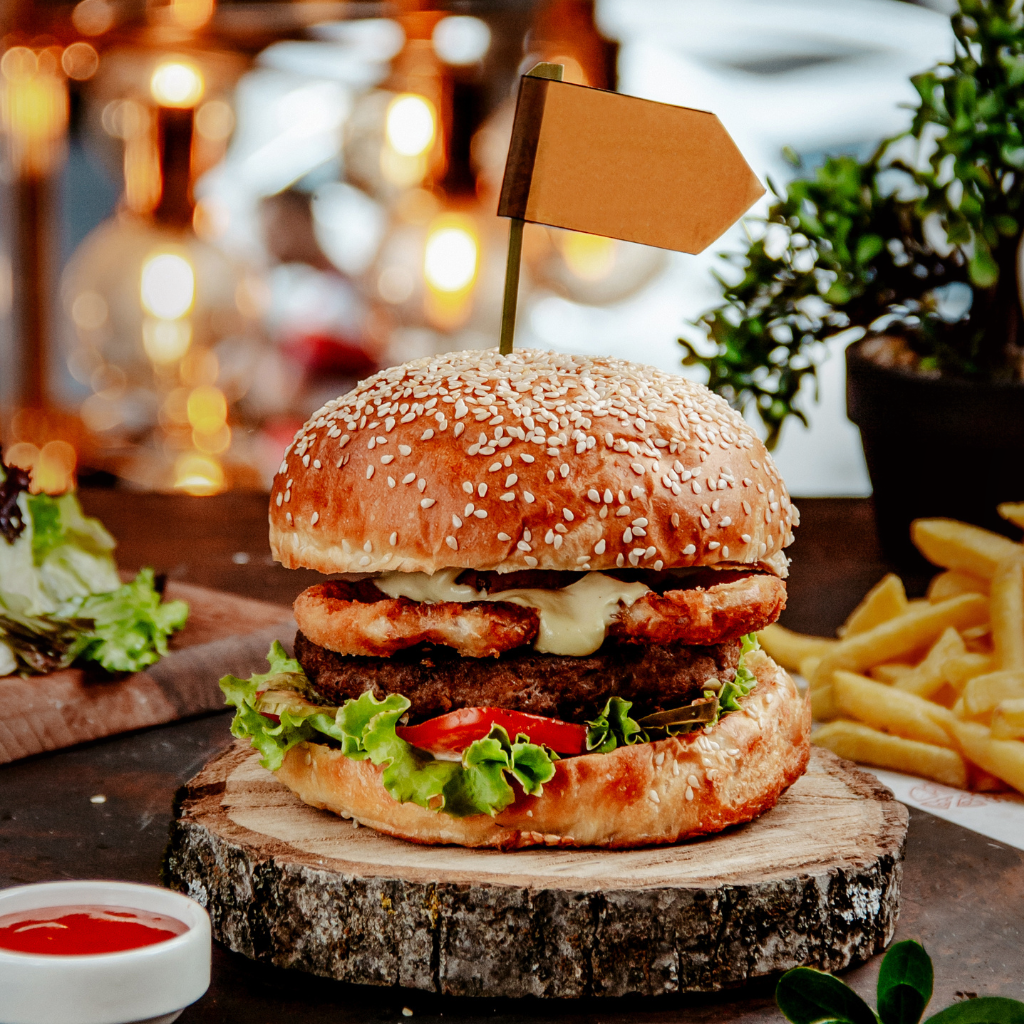 Close-up of a gourmet burger with crispy chicken, lettuce, tomato, and a sesame seed bun, served with fries and dipping sauce.