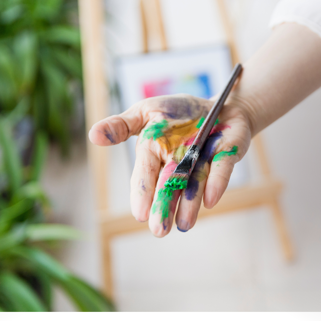 A hand covered in colorful paint holding a paintbrush, representing playful creativity and hands-on interaction.